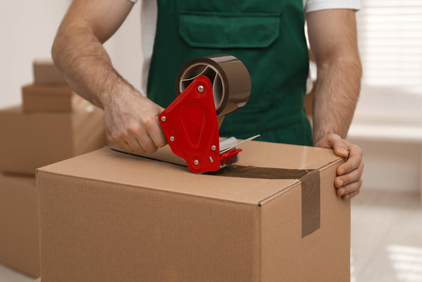 Man packing box with adhesive tape indoors, closeup. Moving serv - Zdjęcie, obraz