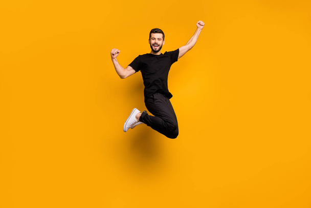 Full body photo of handsome excited guy jumping high raising fists celebrating football team goal supporting fan wear black t-shirt pants isolated yellow color background - Φωτογραφία, εικόνα