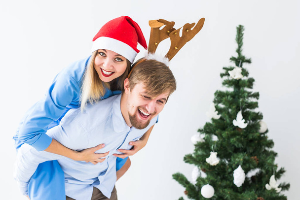 Funny man giving piggyback to his wife while they wearing Santa hats for Christmas holidays at home. - 写真・画像