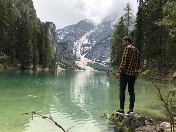 вид ззаду чоловіка мандрівника, що стоїть на березі озера серед гір, вкритих лісом вдень
  - Фото, зображення