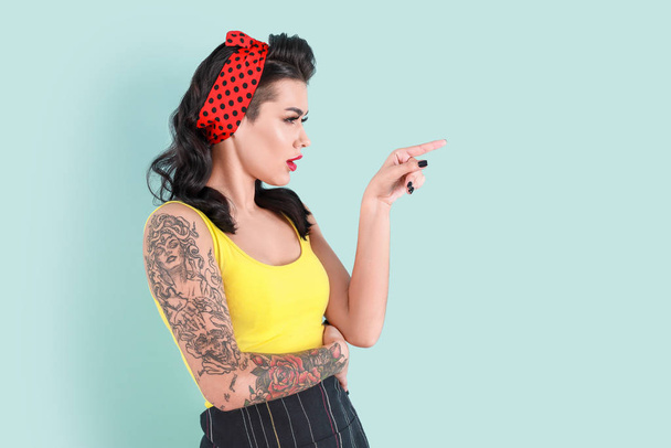 Portrait of beautiful tattooed pin-up woman pointing at something on color background - Photo, Image