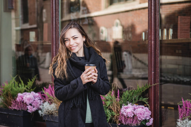 Γυναικείο χέρι με χάρτινο φλιτζάνι καφέ. Γυναίκα κρατώντας κύπελλο στο αστικό φόντο - Φωτογραφία, εικόνα