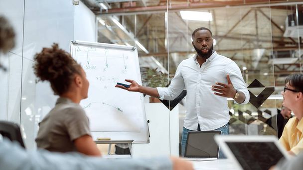 Goede teamleider. Jonge Afro-Amerikaanse zakenman die naar wit schoolbord wijst en zijn collega 's nieuwe projecten uitlegt terwijl hij samenwerkt in het creatieve kantoor - Foto, afbeelding