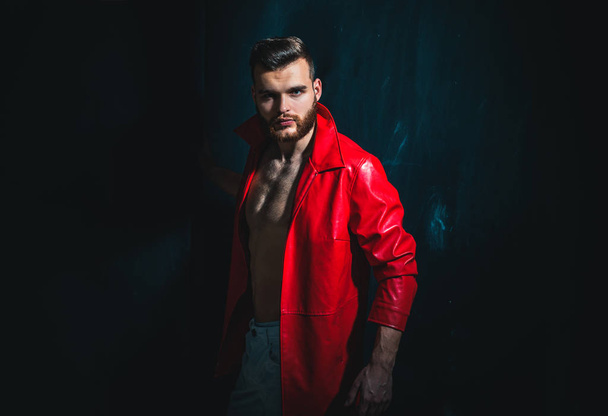 Fashionable man in red jacket is standing on dark background and looking at camera. Portrait of thinking stylish young man. Man with confident face and brutal style. - Valokuva, kuva