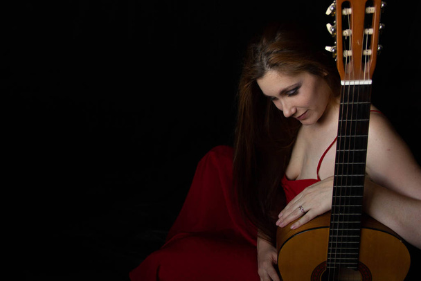 girl in a red dress sits on the floor near the guitar on a black background - Foto, Imagen