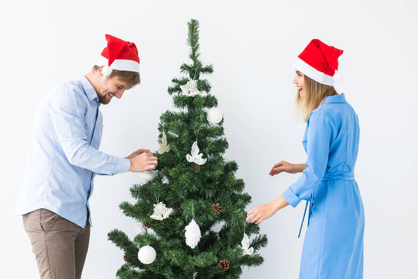 Holidays and festive concept - Young family couple in santa hats decorating the christmas tree - 写真・画像