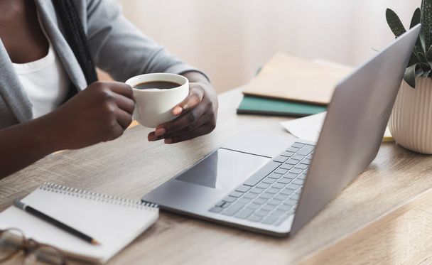 Afro empresaria tomando café mientras trabaja en el ordenador portátil en la oficina
 - Foto, Imagen