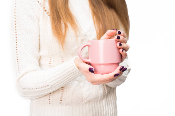 Ένα κορίτσι με ένα λευκό πουλόβερ με σκούρο μανικιούρ κρατά μια κούπα ζεστό ποτό στα χέρια της στο στούντιο. Μεμονωμένα - Φωτογραφία, εικόνα