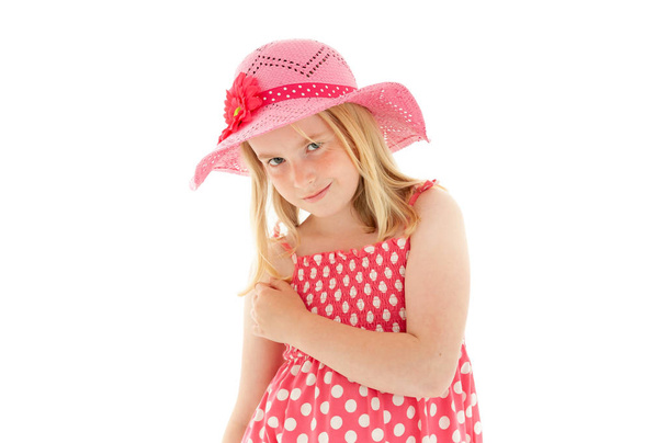 Mooi jong blond meisje met raadselachtige glimlach met grote roze floppy hoed en een polka dot jurk. Geïsoleerd op witte studio achtergrond - Foto, afbeelding