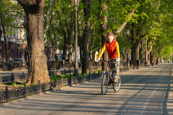 Un niño de 8 años en un chaleco naranja monta una bicicleta en un parque de la ciudad en una soleada mañana de primavera
. - Foto, Imagen