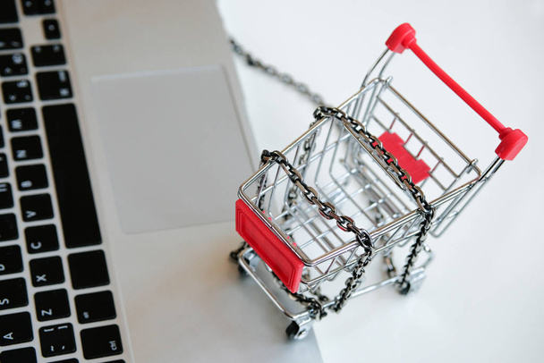 Een grijze laptop op een tafel bij een trolley met een opgerolde ketting uit een supermarkt. Begrip "shopaholic disease". - Foto, afbeelding