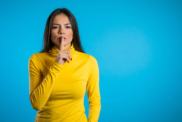 Hispanic beautiful woman holding a finger on her lips over blue background. Gesture of shhh, secret, silence. Close up. Copy space - Zdjęcie, obraz