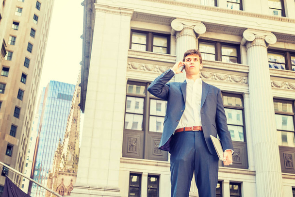 Modern dagelijks leven. Jonge zakenman reist, werkt in New York City, draagt een blauw pak, wit shirt, houdt laptop computer vast, staat op straat buiten kantoorgebouw, praat op mobiele telefoon - Foto, afbeelding