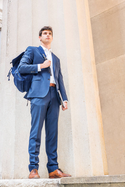 Young businessman street fashion in New York City, wearing blue suit, white shirt, brown leather shoes, shoulder carry bag, holding laptop computer, standing by column outside, looking forward - Fotoğraf, Görsel