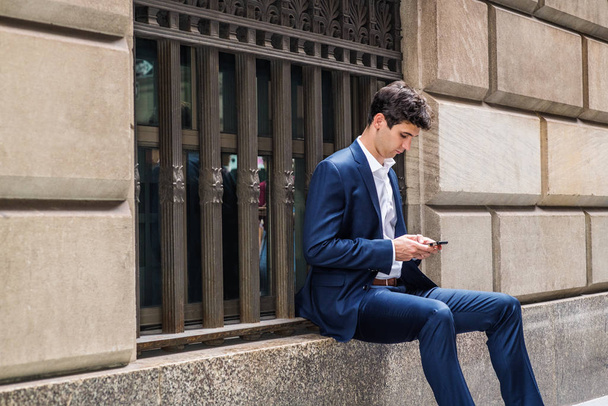 Joven hombre de negocios guapo viajando por la ciudad de Nueva York, vistiendo traje azul, camisa blanca, sentado en un marco de ventana grande de la calle vieja, mirando hacia abajo, leyendo, enviando mensajes de texto en el teléfono celular. Concepto para la vida urbana
 - Foto, imagen