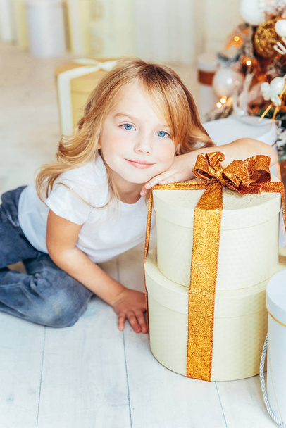 Little girl with gift box near Christmas tree on Christmas eve at home. Young kid in light room with winter decoration. Happy family at home. Christmas New Year december time for celebration concept - Photo, Image