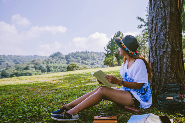 Ασιάτισσα ταξιδεύει στη φύση. Ταξίδι χαλαρώστε. Το κορίτσι που κάθεται και διαβάζει ένα βιβλίο κάτω από το δέντρο. Όμορφο κορίτσι το φθινόπωρο δάσος διαβάζοντας ένα βιβλίο Φύση Εκπαίδευση και Γράψτε μια σημείωση. - Φωτογραφία, εικόνα