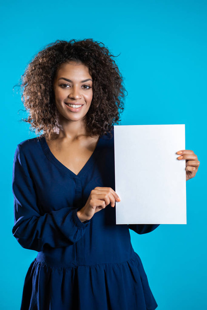 African american woman holding white a4 paper poster. Copy space. Smiling pretty girl with curly hairstyle on blue background. - Photo, Image