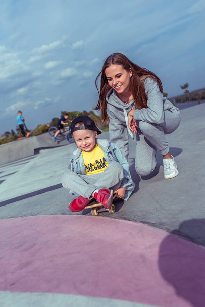 Young mother with little boy son, happy family learns to ride skateboard, summer outdoors, playground. Emotions of delight, joy of happiness and fun. Caring support assistance in training, parenting. - Foto, afbeelding