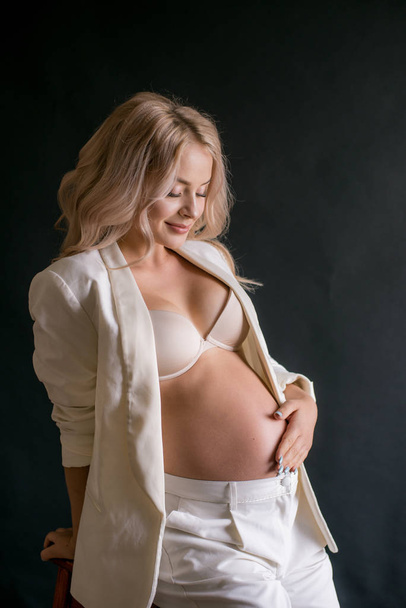 Young beautiful woman with blond long hair pregnant in a white fashionable elegant trouser suit on a black background. Fashion for pregnant women. Happy motherhood - Foto, afbeelding
