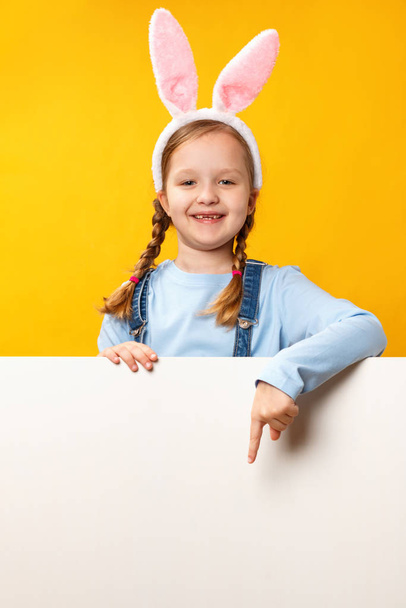 Happy child in rabbit ears shows a finger on a blank banner of space on a yellow background. Easter card - Foto, Bild