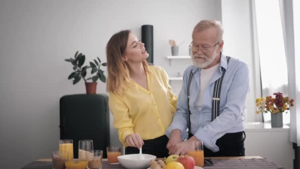 family relationship, happy peppy old man prepares a delicious breakfast in the kitchen with healthy products, mixes with cheerful beautiful adult granddaughter at table background of the kit - Materiał filmowy, wideo