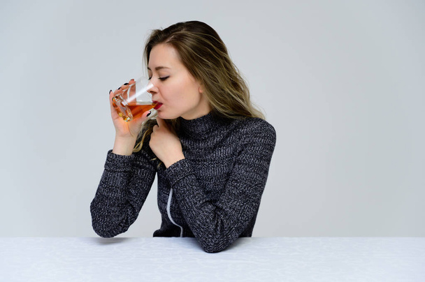 Portrét sedící ženy u stolu, jak mluví s kudrnatými vlasy a emocemi v šedých šatech na bílém pozadí se sklenicí whisky v rukou. Koncept ženského alkoholismu. - Fotografie, Obrázek