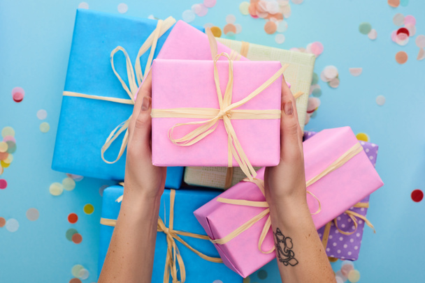 περικοπή άποψη της γυναίκας κρατώντας ροζ κουτί δώρου κοντά πολύχρωμα δώρα κοντά κομφετί στο μπλε  - Φωτογραφία, εικόνα