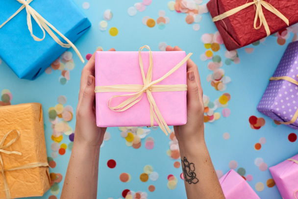 przycięty widok kobiety trzymającej zapakowane różowe pudełko obok kolorowych prezentów w pobliżu confetti na niebiesko  - Zdjęcie, obraz