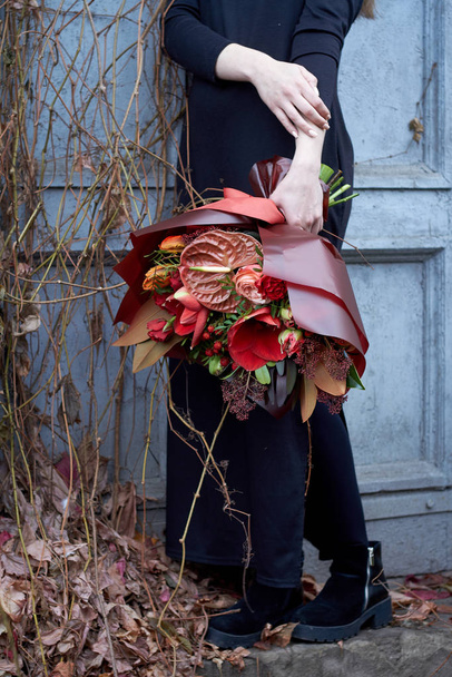 Chica mantiene elegante ramo de otoño en colores rojos en estilo vintage al aire libre en el fondo de las uvas secas y una puerta vieja, enfoque selectivo
 - Foto, Imagen