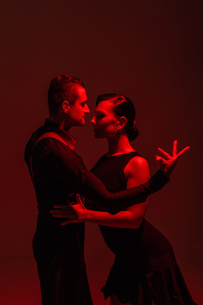leidenschaftliches Tanzpaar in schwarzer Kleidung, das Tango auf dunklem Hintergrund mit roter Beleuchtung aufführt - Foto, Bild