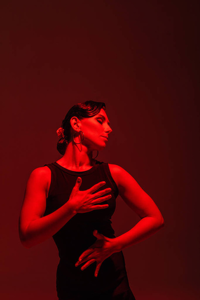 sensual dancer in black dress performing tango on dark background with red lighting - Фото, зображення