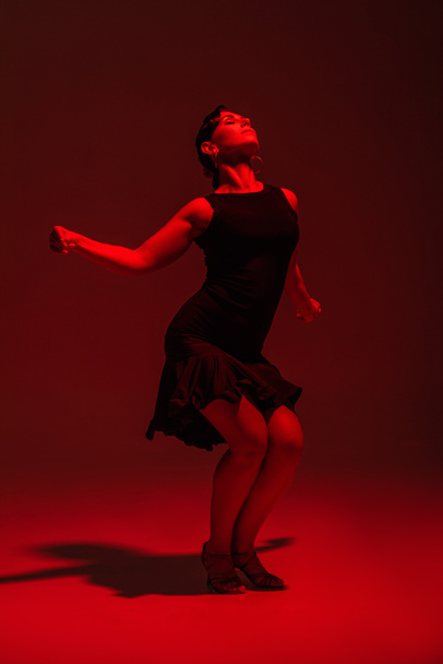 sinnliche Tänzerin im schwarzen Kleid, die Tango auf dunklem Hintergrund mit roter Beleuchtung performt - Foto, Bild