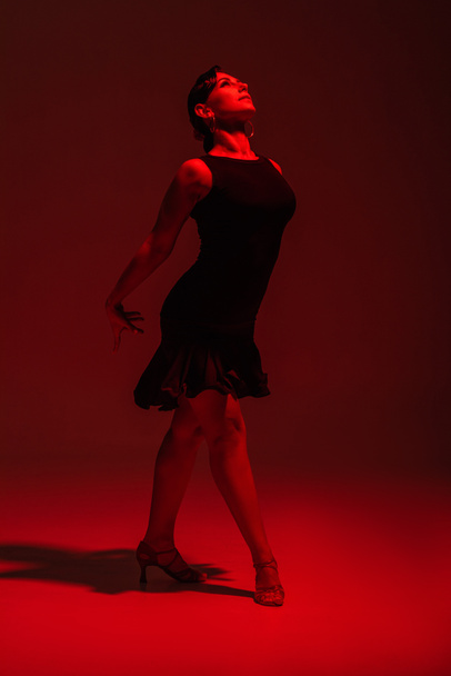 beautiful dancer in black dress performing tango on dark background with red lighting - Фото, изображение