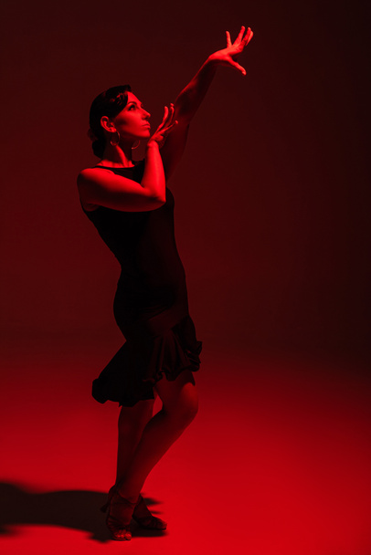 beautiful dancer in black dress performing tango on dark background with red illumination - Foto, imagen