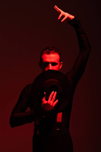 passionate dancer looking at camera and holding hat near face while performing tango on dark background with red illumination - Foto, imagen