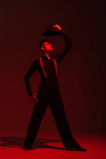 elegante bailarina sosteniendo sombrero sobre la cabeza mientras realiza tango sobre fondo oscuro con iluminación roja
 - Foto, imagen