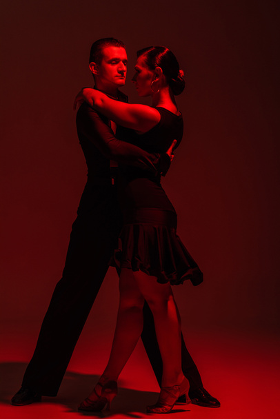 elegant couple of dancers performing tango on dark background with red illumination - Photo, Image