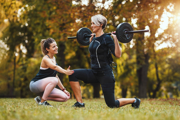 Dojrzała kobieta wykonuje ćwiczenia z osobistym trenerem w parku, ubrana w czarny garnitur z elektronicznym symulatorem Ems, aby pobudzić jej mięśnie. - Zdjęcie, obraz