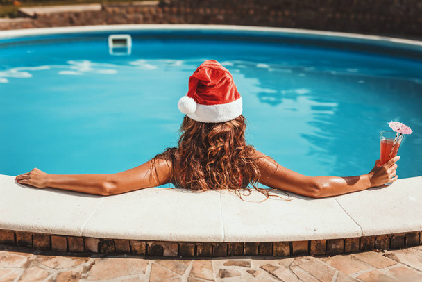 Joven hermosa mujer cerca de la piscina en el sombrero de Santa Claus, está celebrando el Año Nuevo y la Navidad en el país caliente con una copa de cóctel
. - Foto, Imagen
