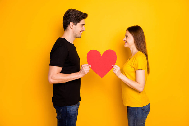 Photo side profile of two beloved cheerful charming cute nice couple wearing jeans denim black t-shirt looking into each other eyes holding red heart isolated vivid color background - Φωτογραφία, εικόνα