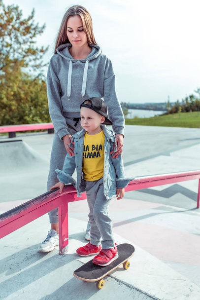Young mother teaches to ride a skateboard, son of a little boy, summer day. Autumnal clothes, casual clothes. Help and support in parenting. Caring for children. - Foto, Bild