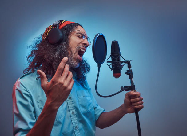 Africano cantante rastafari masculino usando una camisa azul y gorro emocionalmente escribir canción en el estudio de grabación
. - Foto, imagen