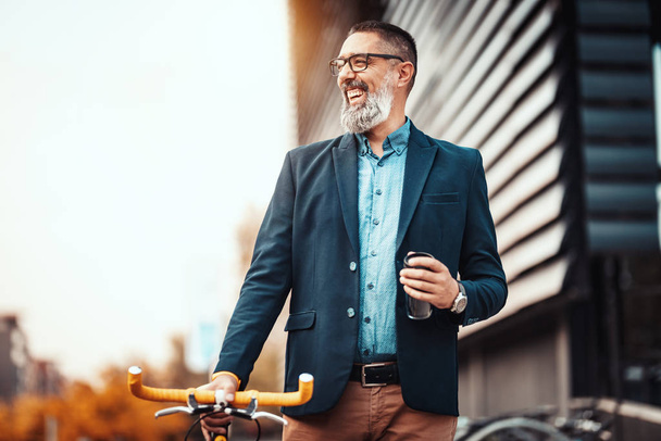 Exitoso hombre de negocios guapo de mediana edad con una taza de café para ir y con bicicleta a su lado, camina frente al distrito de oficinas
. - Foto, imagen