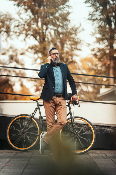 Ένας όμορφος μεσήλικας πάει στη δουλειά με ποδήλατο και μιλάει στο smartphone.. - Φωτογραφία, εικόνα