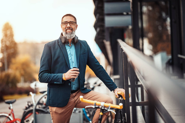Успішний бізнесмен середнього віку з чашкою кави, щоб піти і з велосипедом поруч з ним стоїть перед офісним районом
. - Фото, зображення
