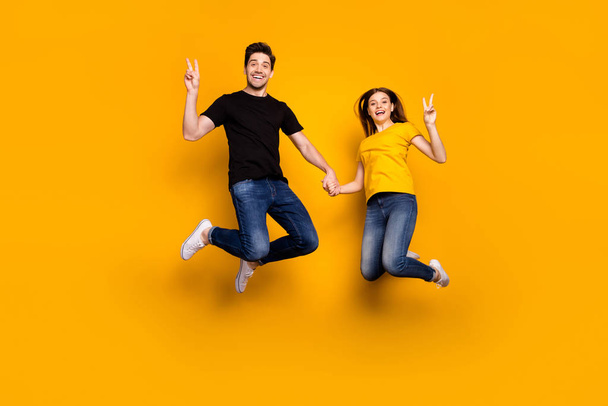 Full body photo of crazy guy lady couple jumping high holding arms showing v-sign symbols saying hi wear casual jeans black t-shirts isolated yellow color background - Photo, image