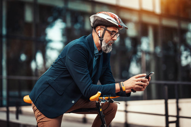 Casual handsome middle-aged businessman is going to work by bicycle. He is standing on the bike and using smart phone in front of office district. - Photo, Image