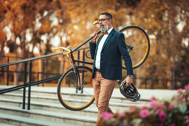 Ένας όμορφος μεσήλικας επιχειρηματίας θα πάει στο γραφείο με ποδήλατο. Κουβαλάει ένα ποδήλατο στον ώμο του πάνω από τις σκάλες.. - Φωτογραφία, εικόνα