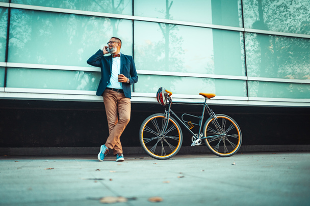Un bel homme d'affaires d'âge moyen se tient à côté de son vélo et utilise un téléphone intelligent devant le quartier des bureaux
 - Photo, image